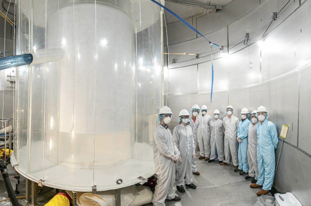 Ein Team von Wissenschaftlern in Ganzkörperschutzanzügen steht in einer Reihe neben einem Gerät, das konzentrische Zylinder zu sein scheint, mit einem transparenten Äußeren und einem undurchsichtigen Inneren, das sich vom Boden bis zur Decke erstreckt.