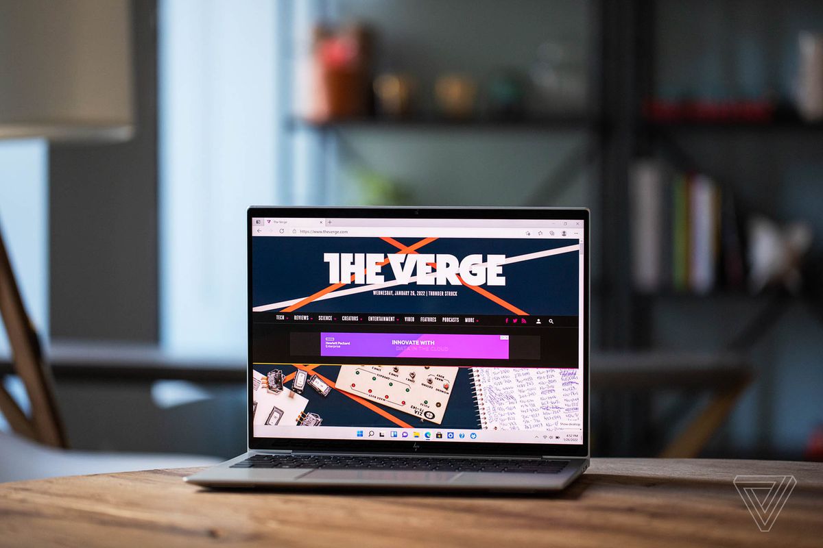 HP Elite Dragonfly G3 an einem Holztisch mit Bücherregal im Hintergrund, offen, leicht nach links geneigt.  Auf dem Bildschirm wird die Startseite von The Verge angezeigt.