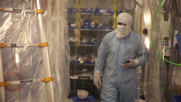 Ein Laborant trägt einen Vollschutzanzug mit Kopf- und Gesichtsbedeckung in einem mit Plastikplanen abgedeckten Raum.